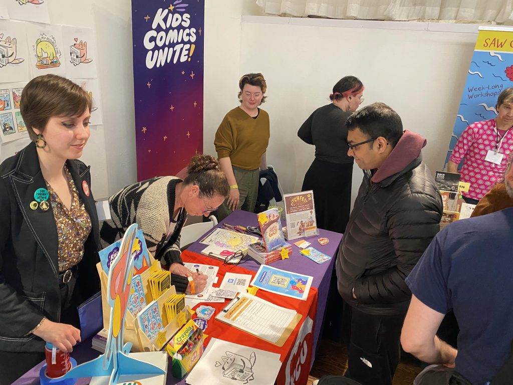 Let's Go! contributor Hollie Michaels signs a copy of the anthology at KCU's table at MoCCA Fest 2026. 